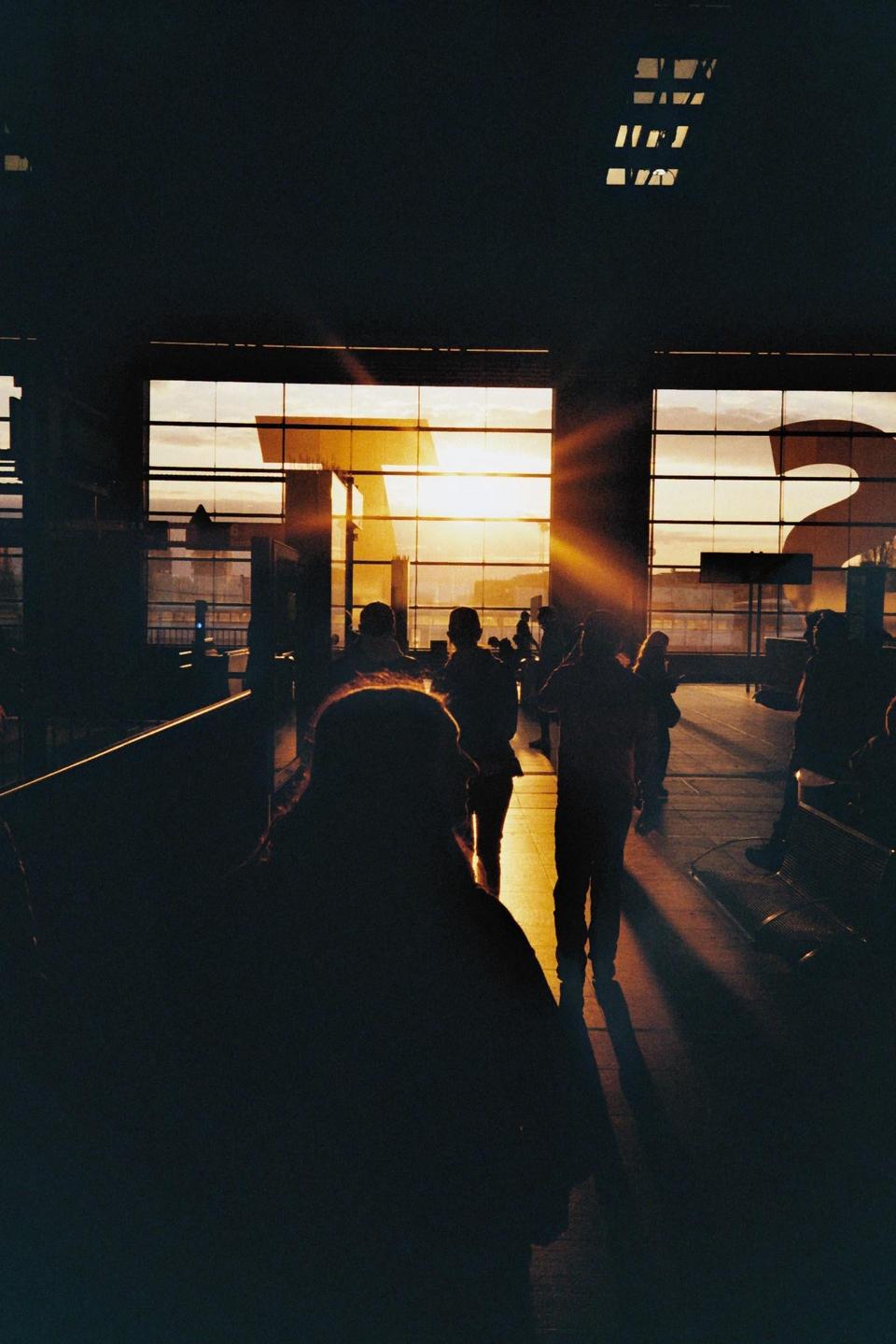 Silhouetted people stand in a building with large windows looking out onto a sunset.