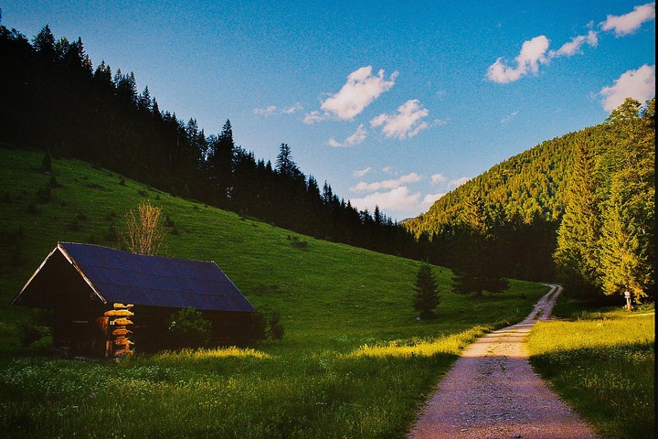 A dirt road leads through a green valley with a small wooden cabin on the left and a forest on the hillsides under a blue sky with clouds.