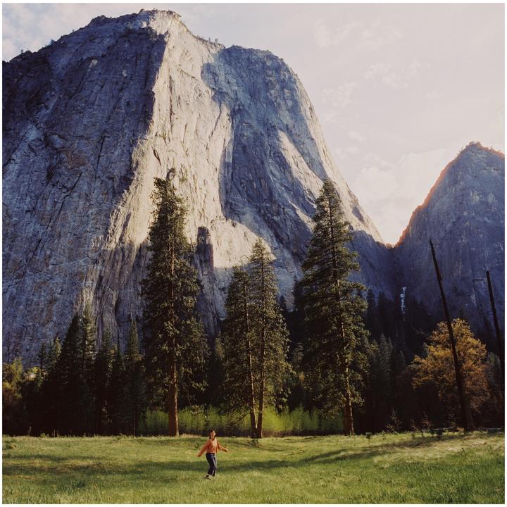 A woman stands in a grassy field with tall pine trees and a large mountain in the background.