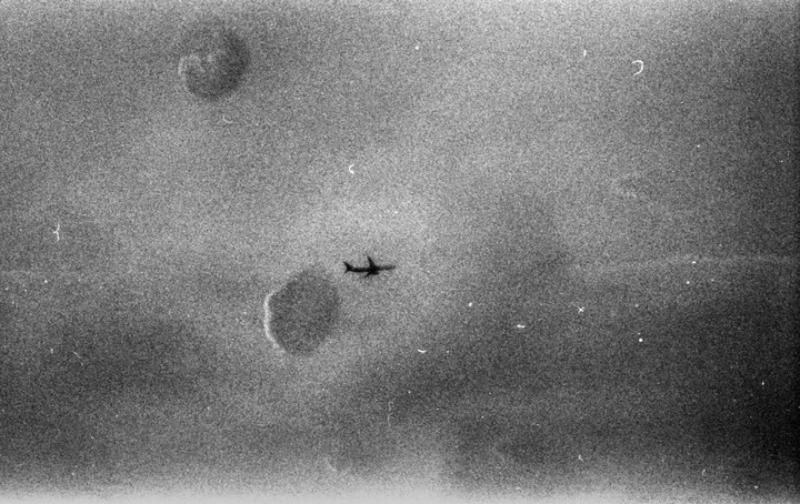 A grainy, black and white image shows a small airplane flying across a cloudy sky.
