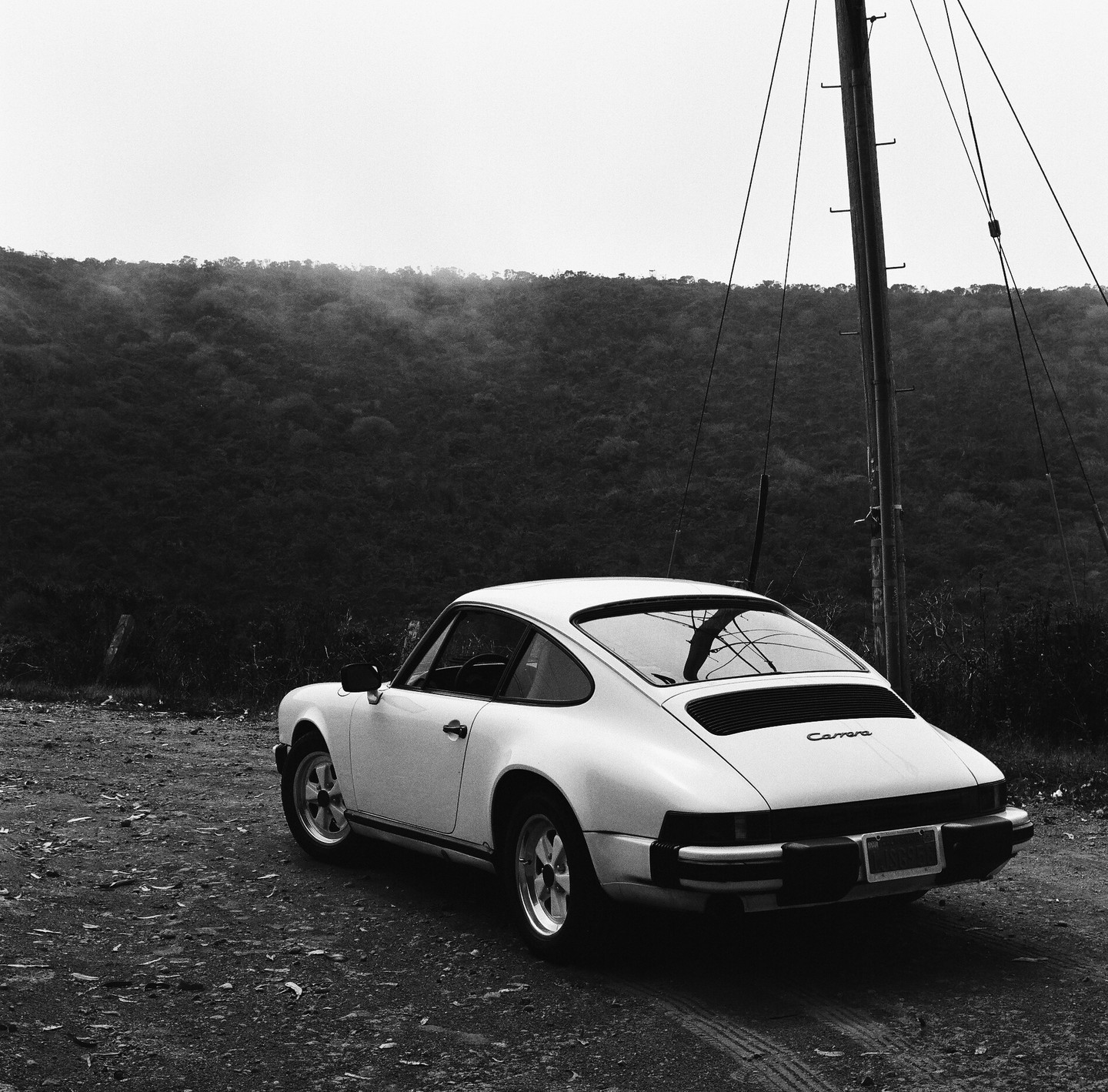 A white Porsche is parked on a dirt road next to a utility pole with a forested hill in the background.