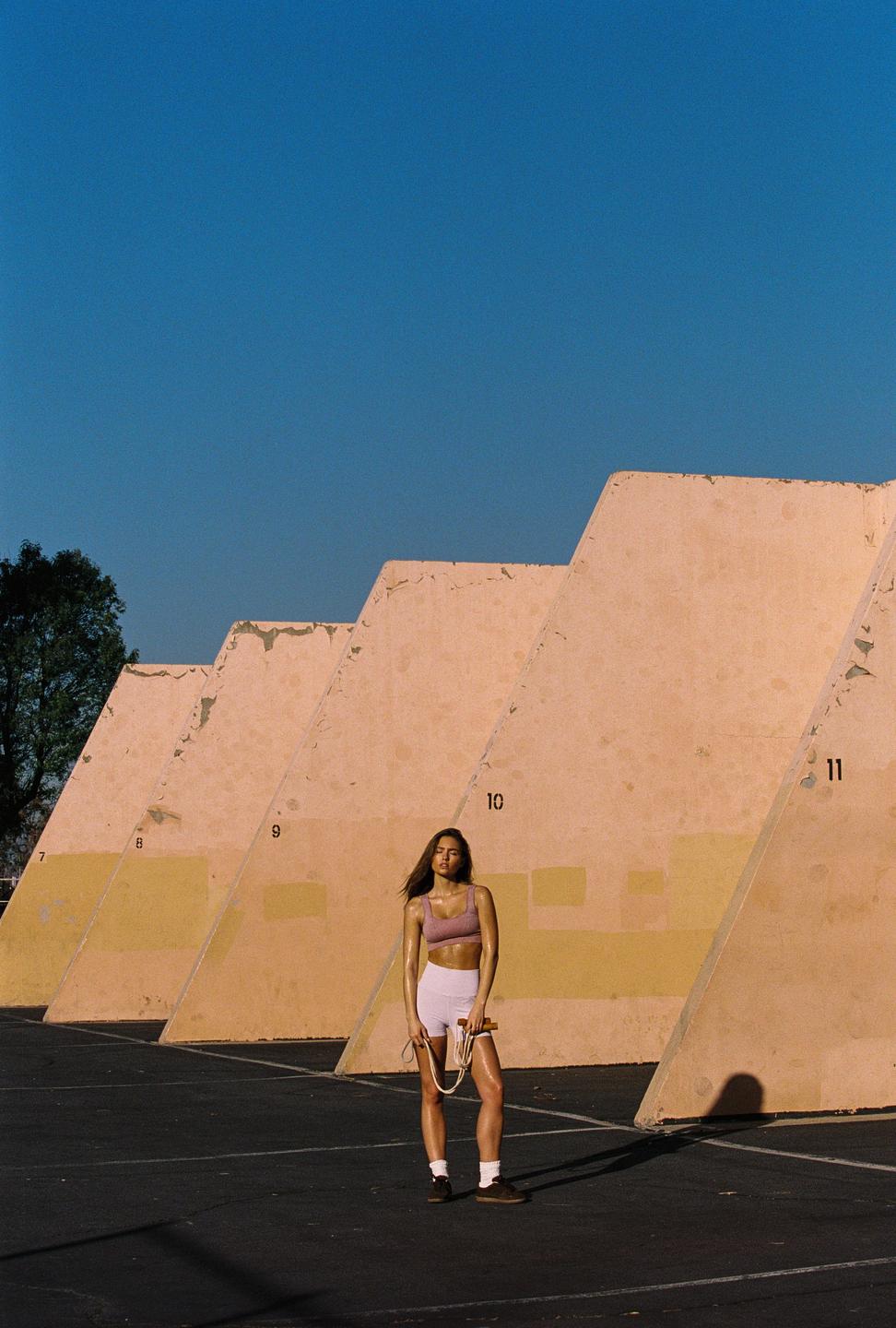 A woman in athletic wear stands on an asphalt court holding a jump rope with a series of numbered concrete walls behind her.
