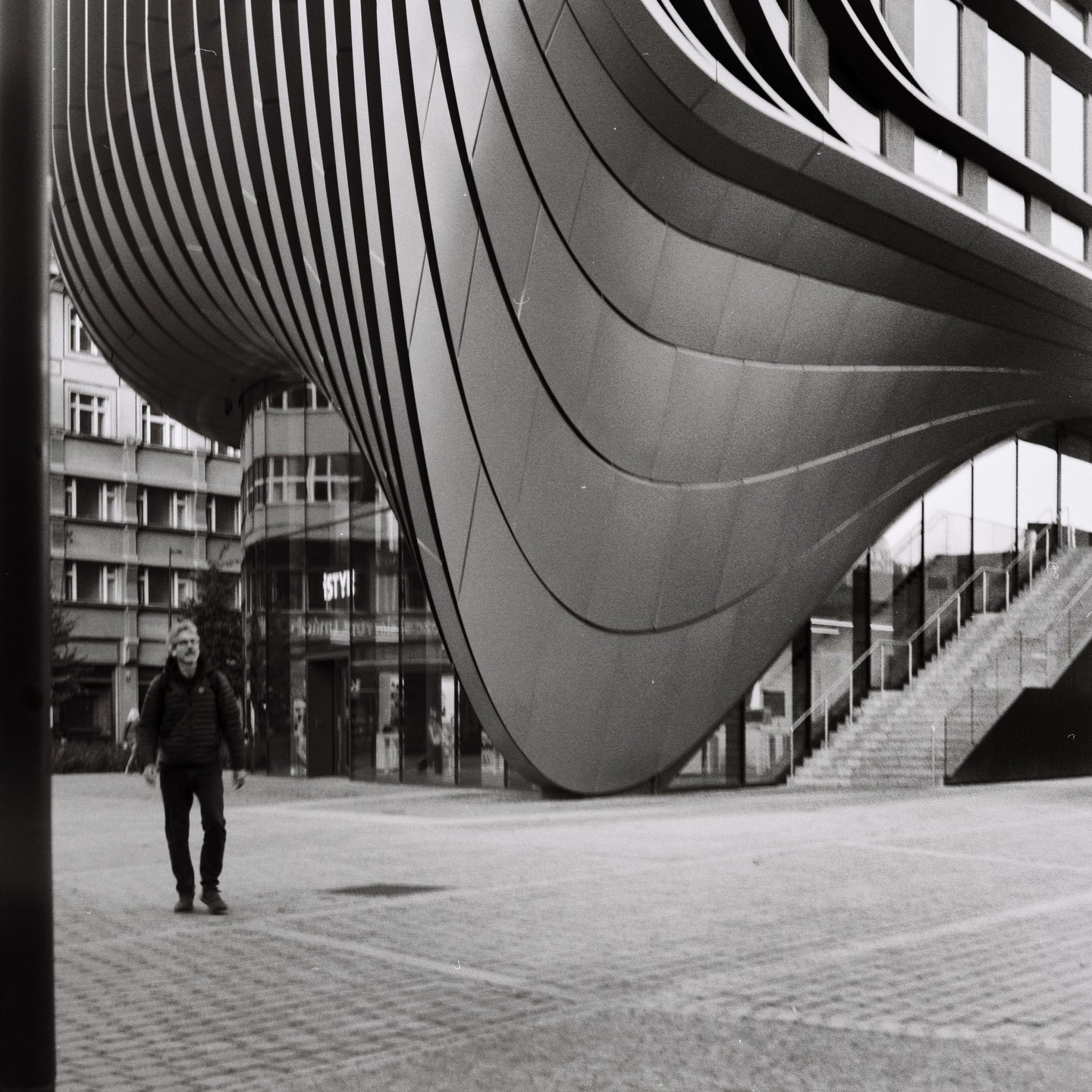 A man walks across a paved plaza next to a modern building with a curved, ribbed facade and a staircase.