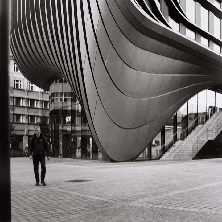 A man walks across a paved plaza next to a modern building with a curved, ribbed facade and a staircase.