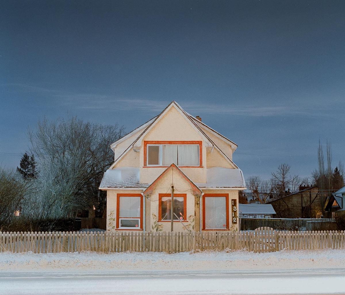A cream-colored house with orange trim and a picket fence stands in a snowy landscape under a dark sky.