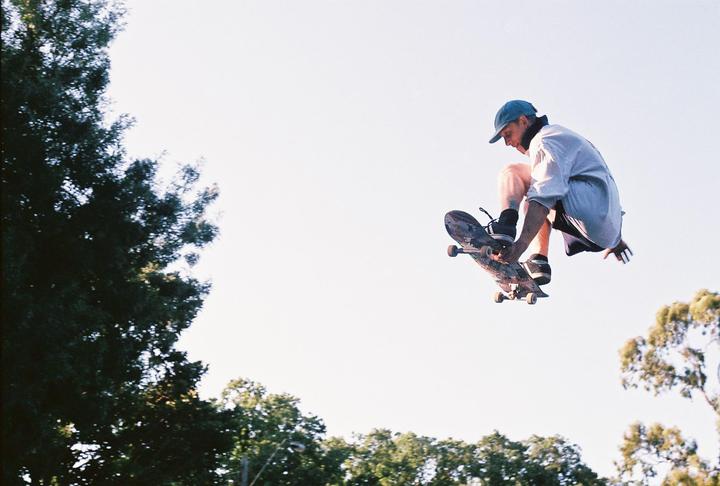 A man in a blue cap and white shirt is captured mid-air on a skateboard against a bright sky with trees in the background.