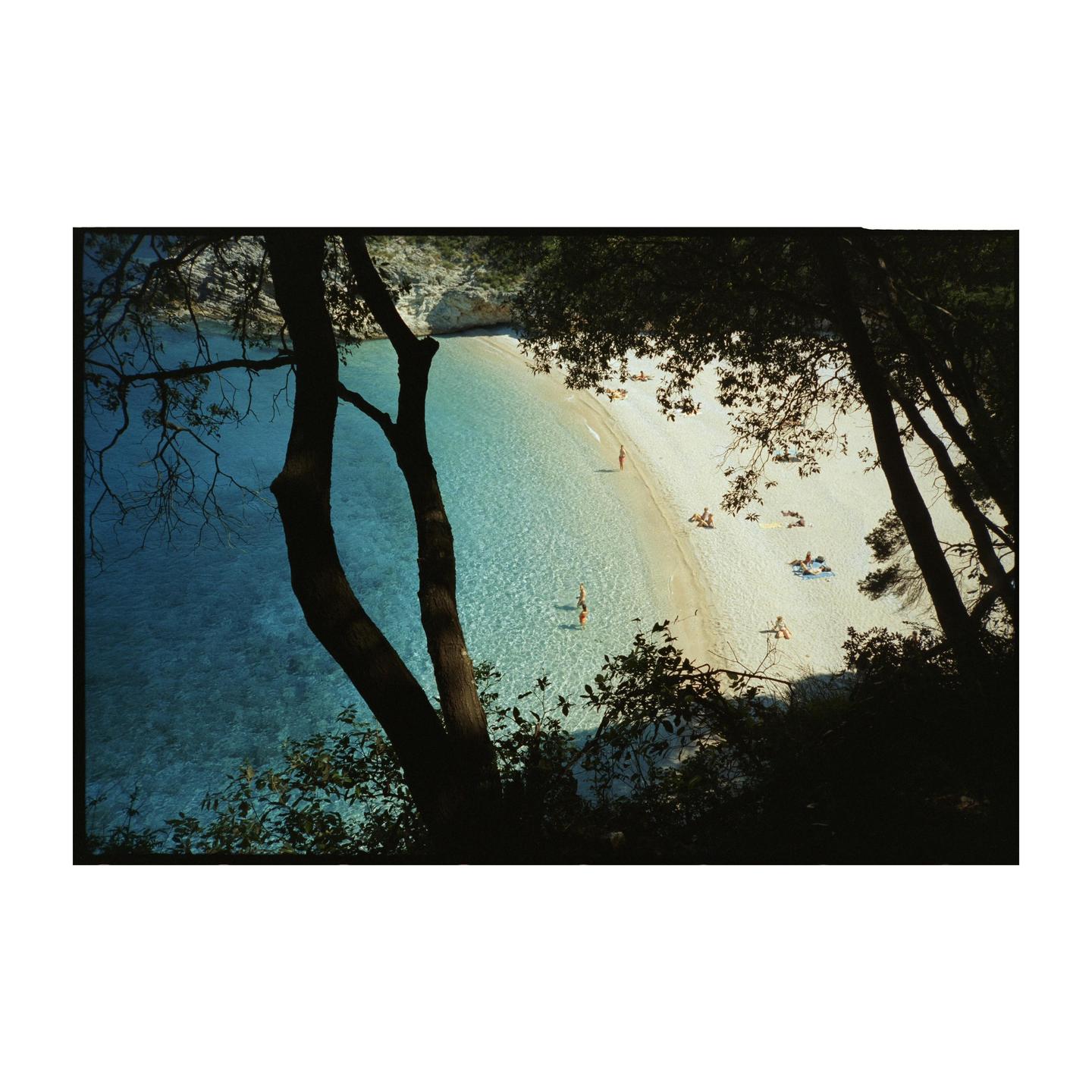 A high-angle view through trees shows a white sand beach with people sunbathing and swimming in clear blue water.