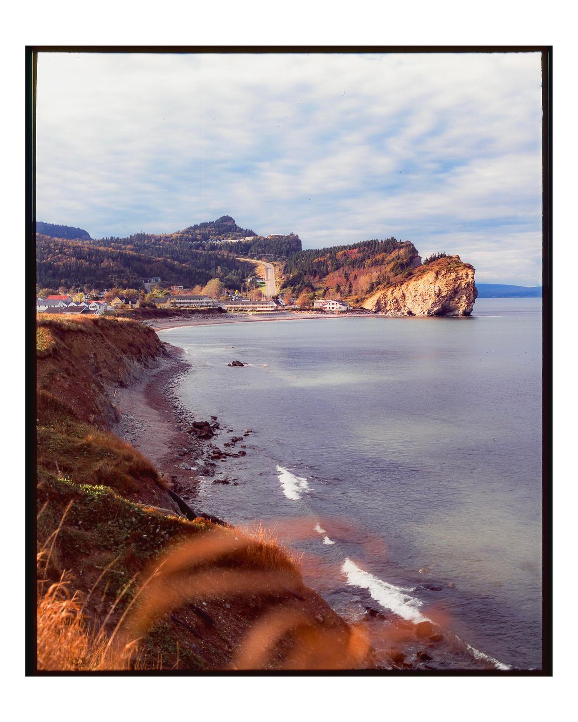 A coastal town with a road winding through hills and a large rock formation extending into the water is visible under a cloudy sky.