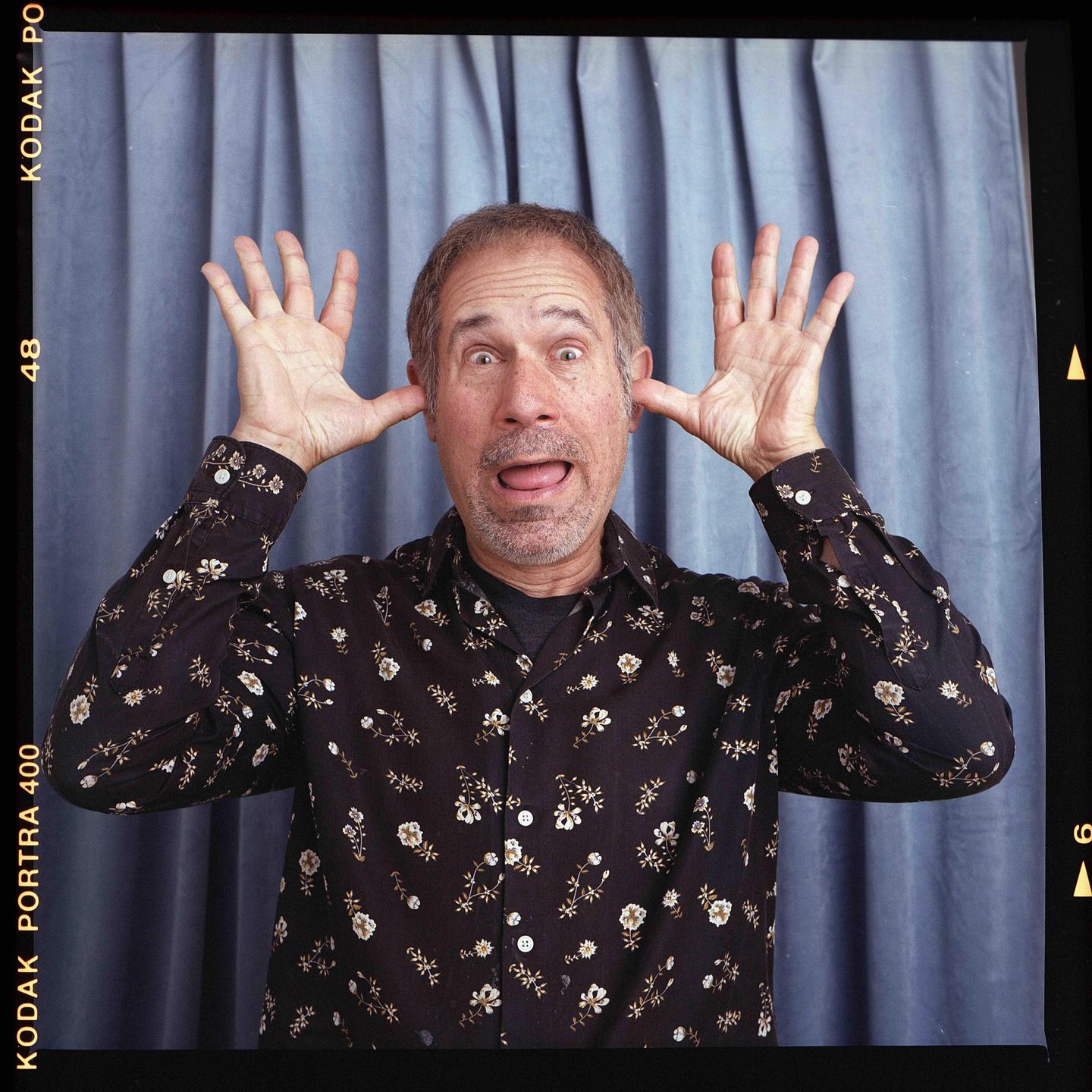A man with a floral shirt makes a silly face with his tongue out and hands by his ears.