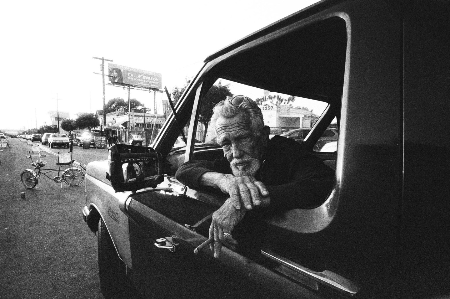 A man with a beard and sunglasses on his head leans out of a truck window, holding a cigar, with a bicycle and street in the background.