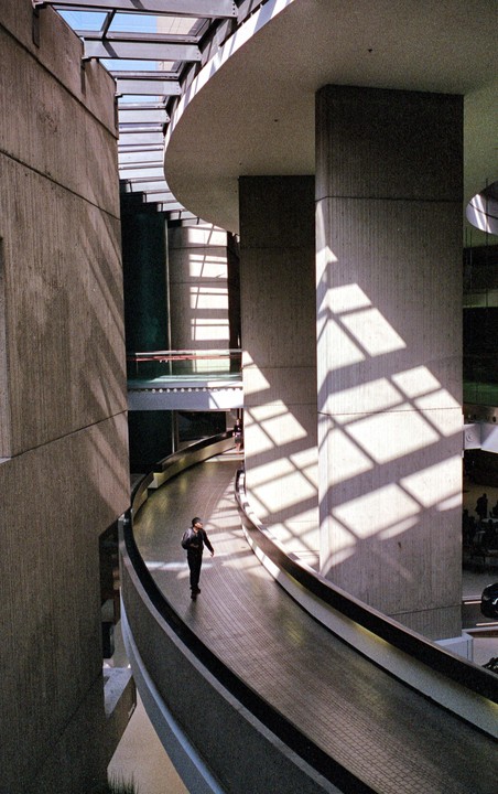 A man walks along a curved walkway inside a brutalist building with strong shadows cast from overhead windows.