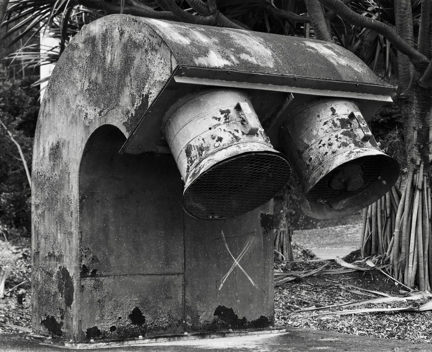 A large, weathered metal structure with two circular vents and an "X" drawn on its side stands among palm trees.