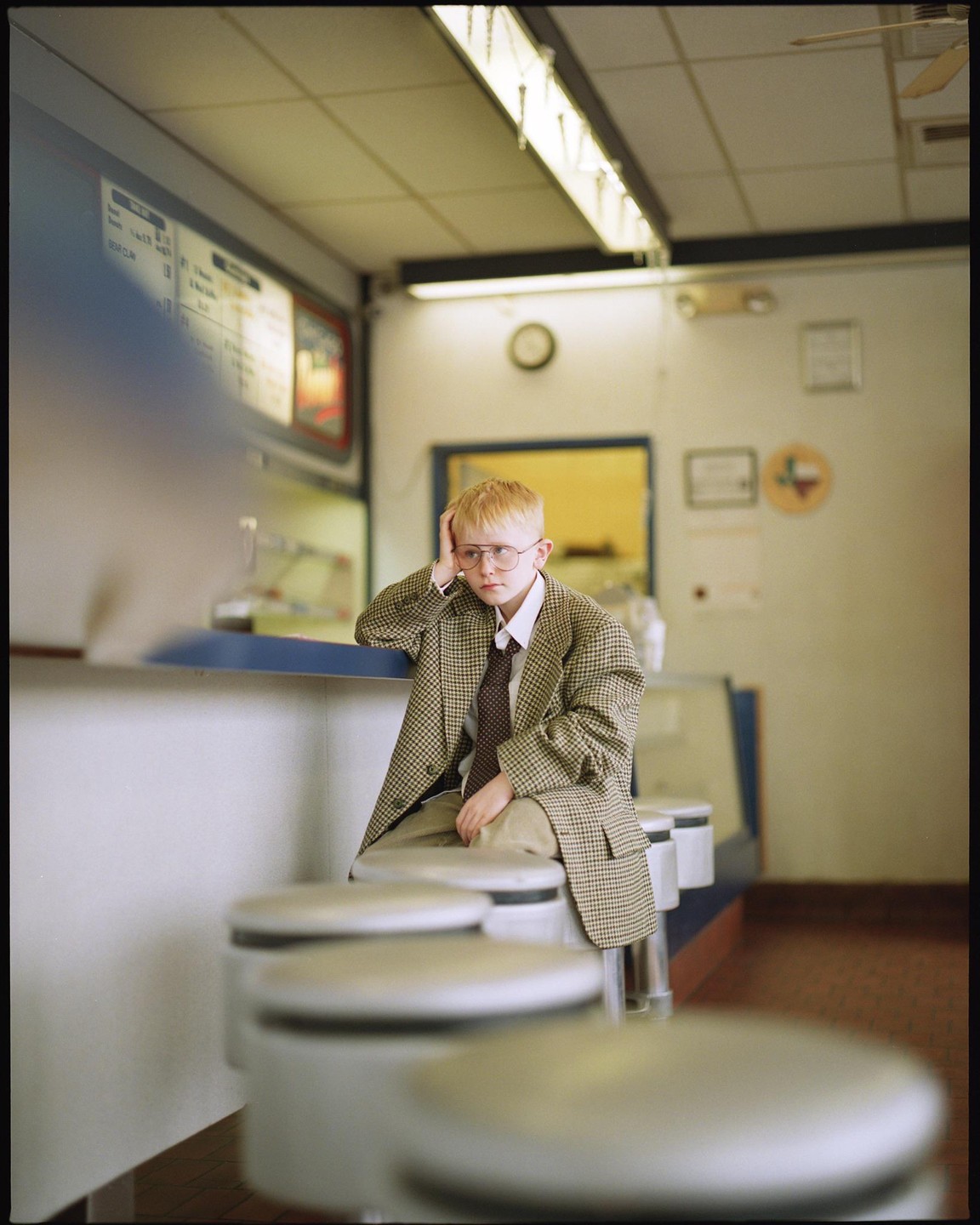 A boy in an oversized suit and glasses sits at a diner counter, resting his head on his hand.