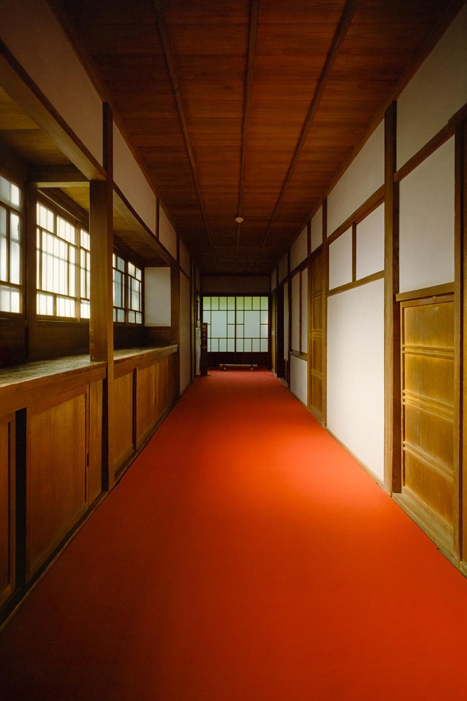 A long, narrow hallway with a red carpet leads to a shoji screen at the end, flanked by wooden walls and windows.