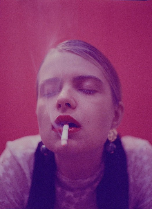 A woman with red lipstick and closed eyes smokes a cigarette, with smoke rising from her mouth.