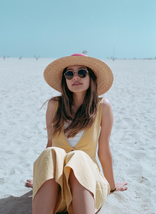 A woman wearing a straw hat and sunglasses sits on a sandy beach.