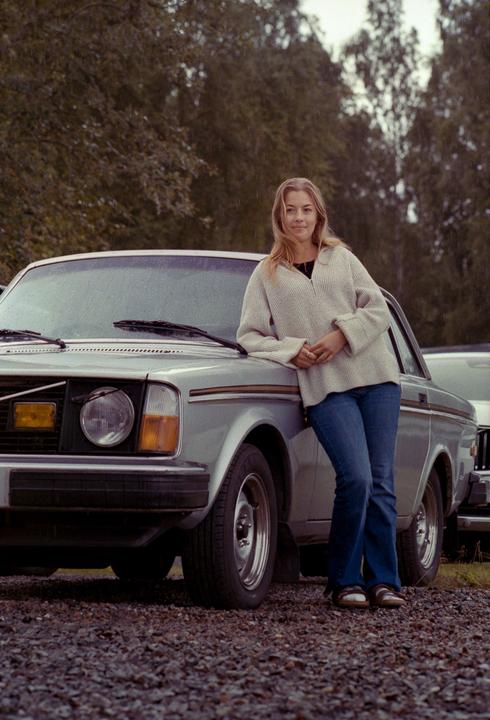 A woman in a sweater and jeans leans against a silver car on a gravel surface with trees in the background.