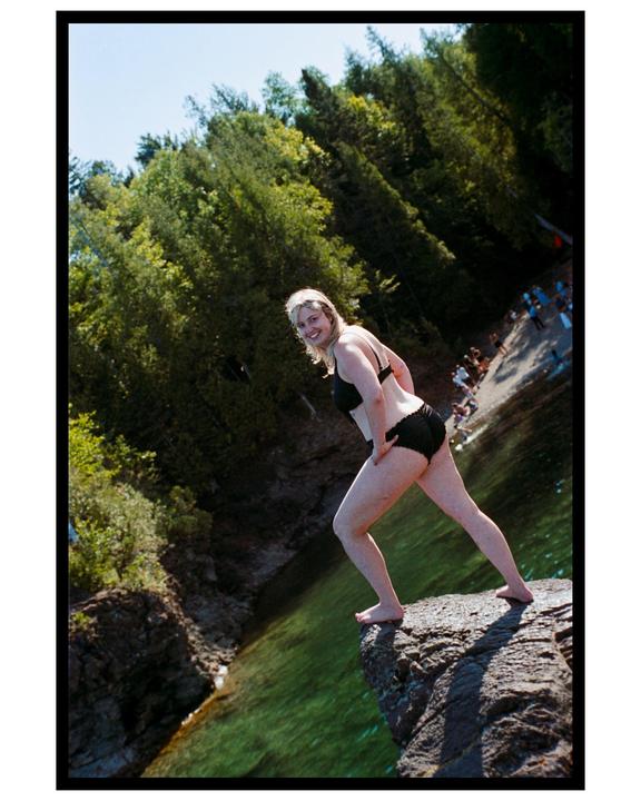 A woman in a black bikini stands on a rock cliff overlooking clear green water and a distant beach with people.