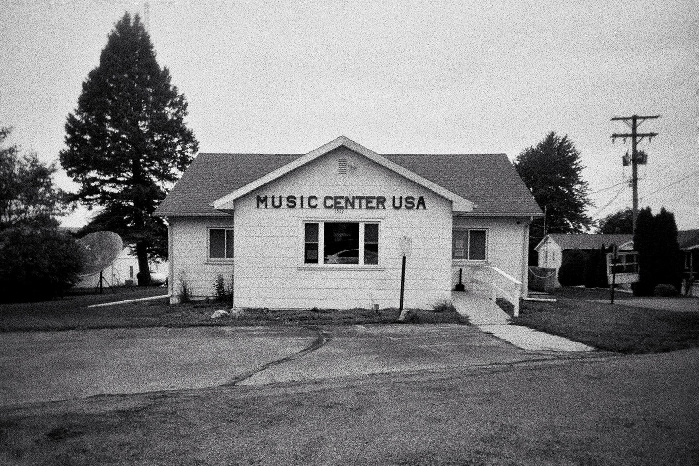 A small building with "MUSIC CENTER USA" written on its facade stands in a grassy area with a paved driveway.