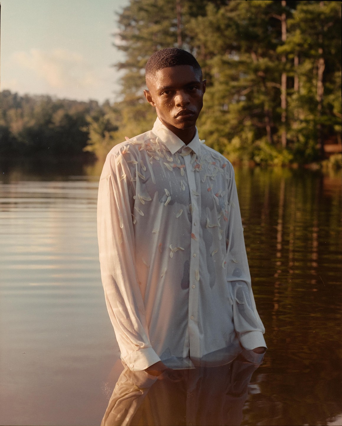 A man stands in a body of water wearing a wet white shirt with petal-like embellishments.