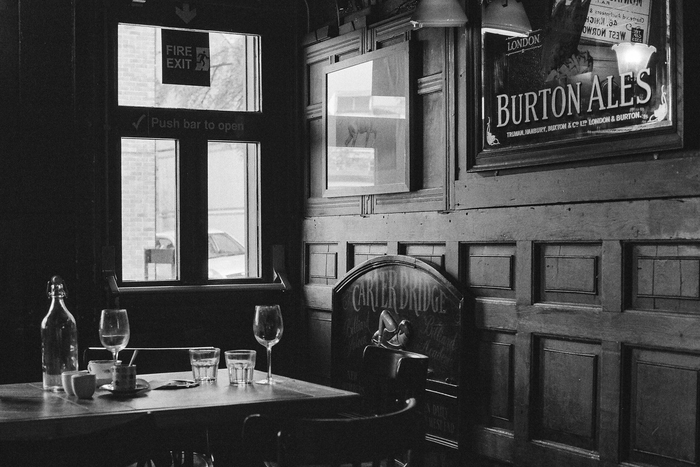 A table with glasses and a bottle sits next to a window in a dimly lit pub with wood paneling and a "Burton Ales" sign.