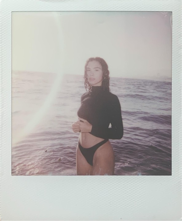 A woman with wet, curly hair stands in the ocean, wearing a black long-sleeve top and black bikini bottom, with sunlight reflecting on the water.