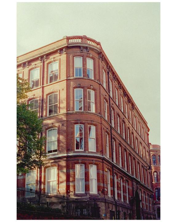 A large brick building with many windows stands at a street corner, with sunlight casting shadows on its facade.