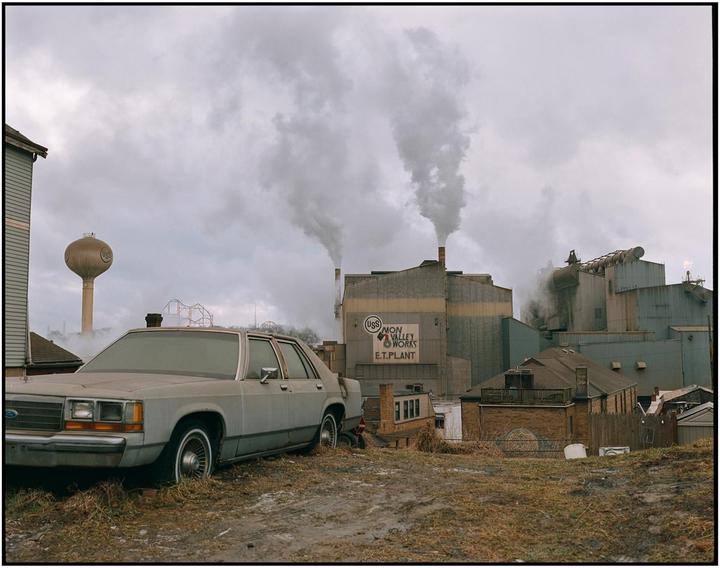 A gray car is parked on a dirt patch with an industrial plant emitting smoke in the background under a cloudy sky.