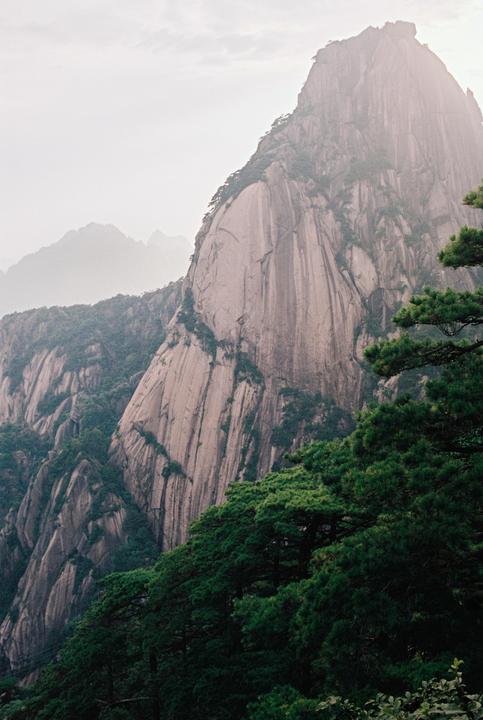 A large, rocky mountain with green trees in the foreground rises into a hazy sky.