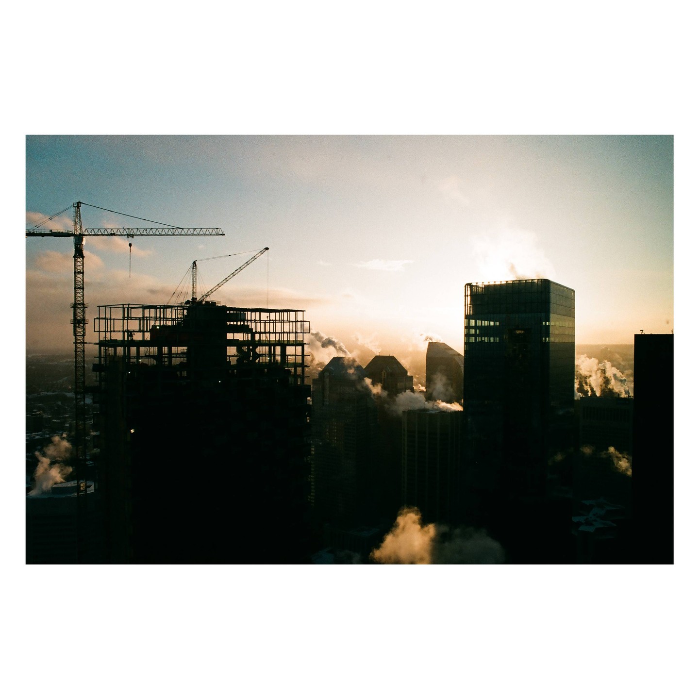A city skyline with construction cranes and buildings emitting steam at sunset.