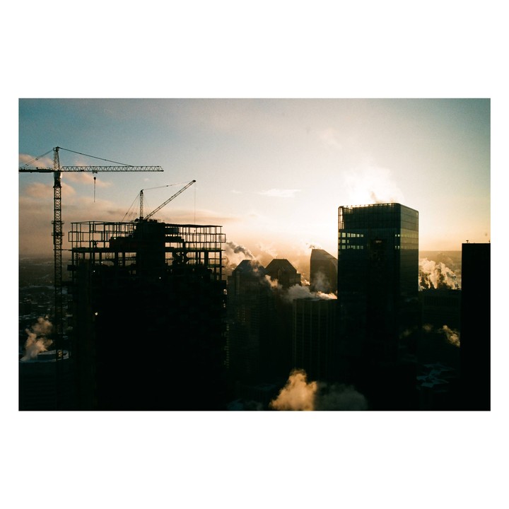 A city skyline with construction cranes and buildings emitting steam at sunset.