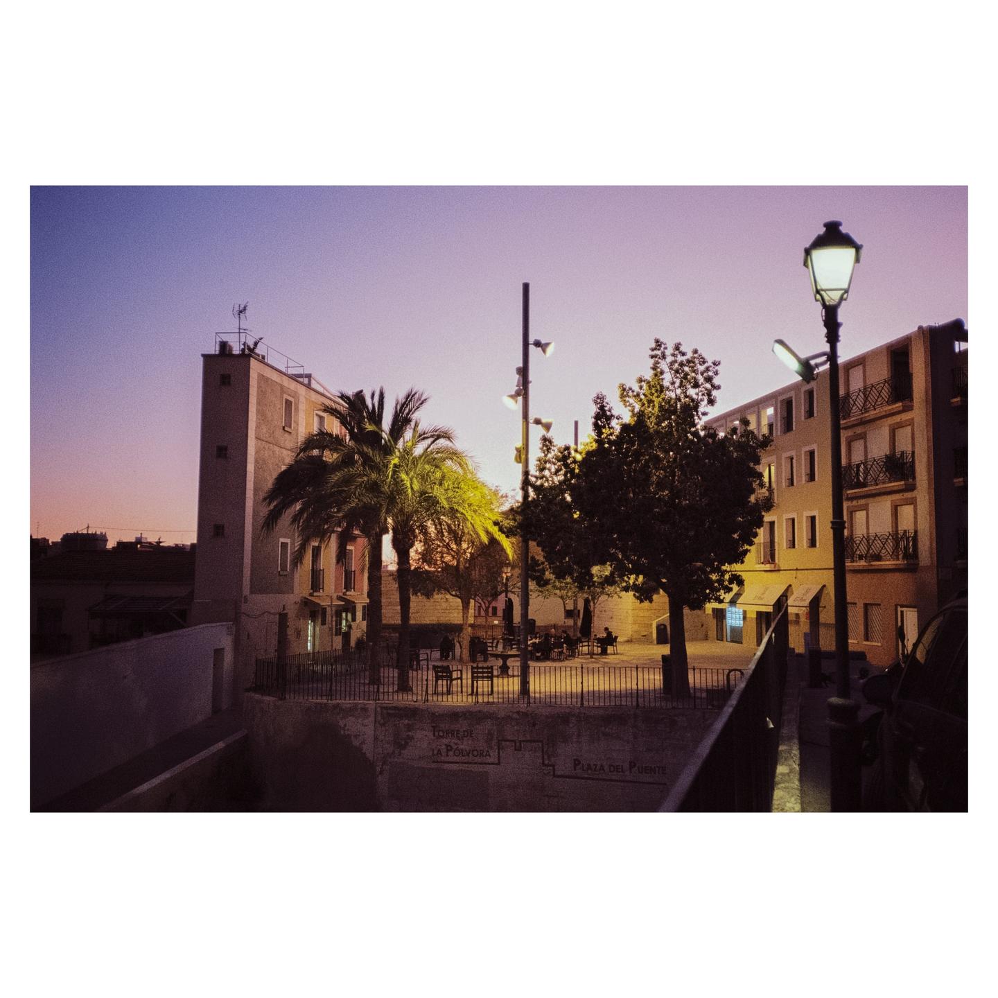 A town square with palm trees and buildings is illuminated by streetlights under a purple sky at dusk.
