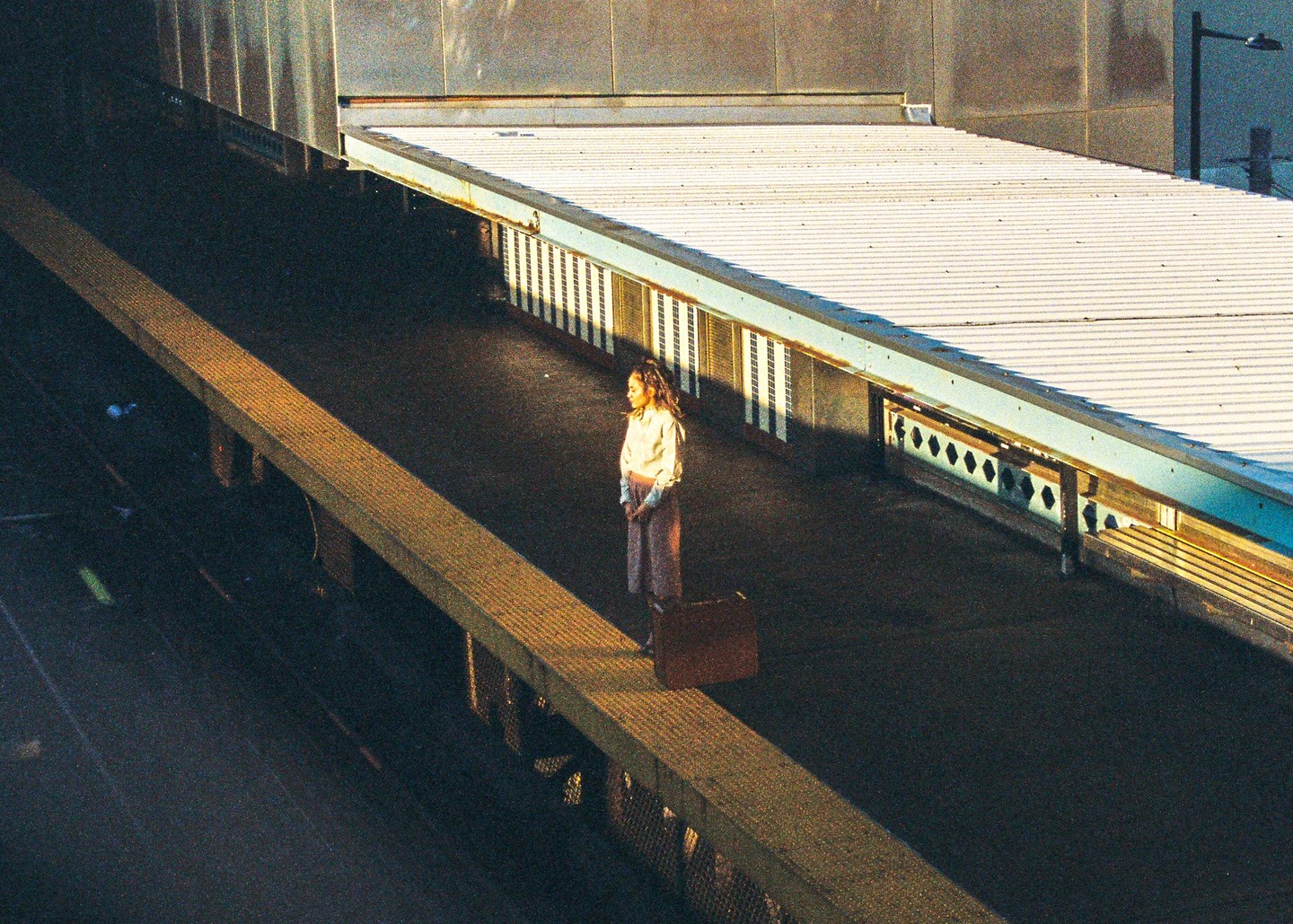 A woman stands on a train platform next to a suitcase, illuminated by sunlight.