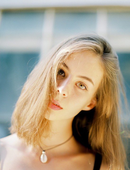 A young woman with long blonde hair and a necklace looks directly at the viewer.