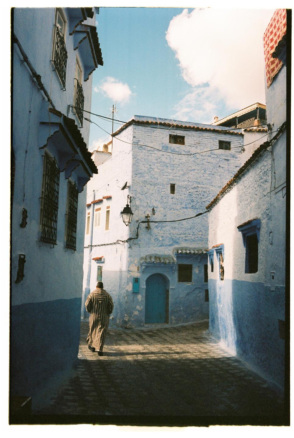 A man in a striped robe walks down a narrow street lined with blue buildings under a cloudy sky.
