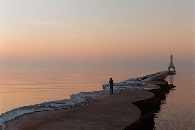 A person stands on a pier leading to a lighthouse at sunset.