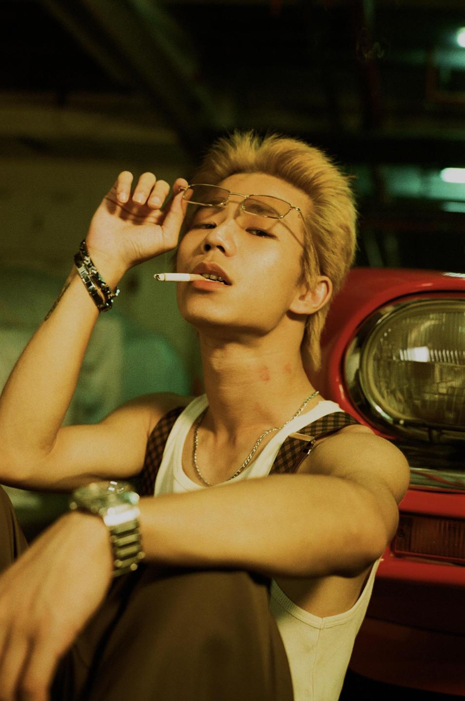 A man with bleached hair and sunglasses holds a cigarette in his mouth while leaning against a red car.