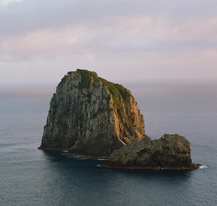 A large rock formation covered in green foliage rises from the ocean under a cloudy sky.