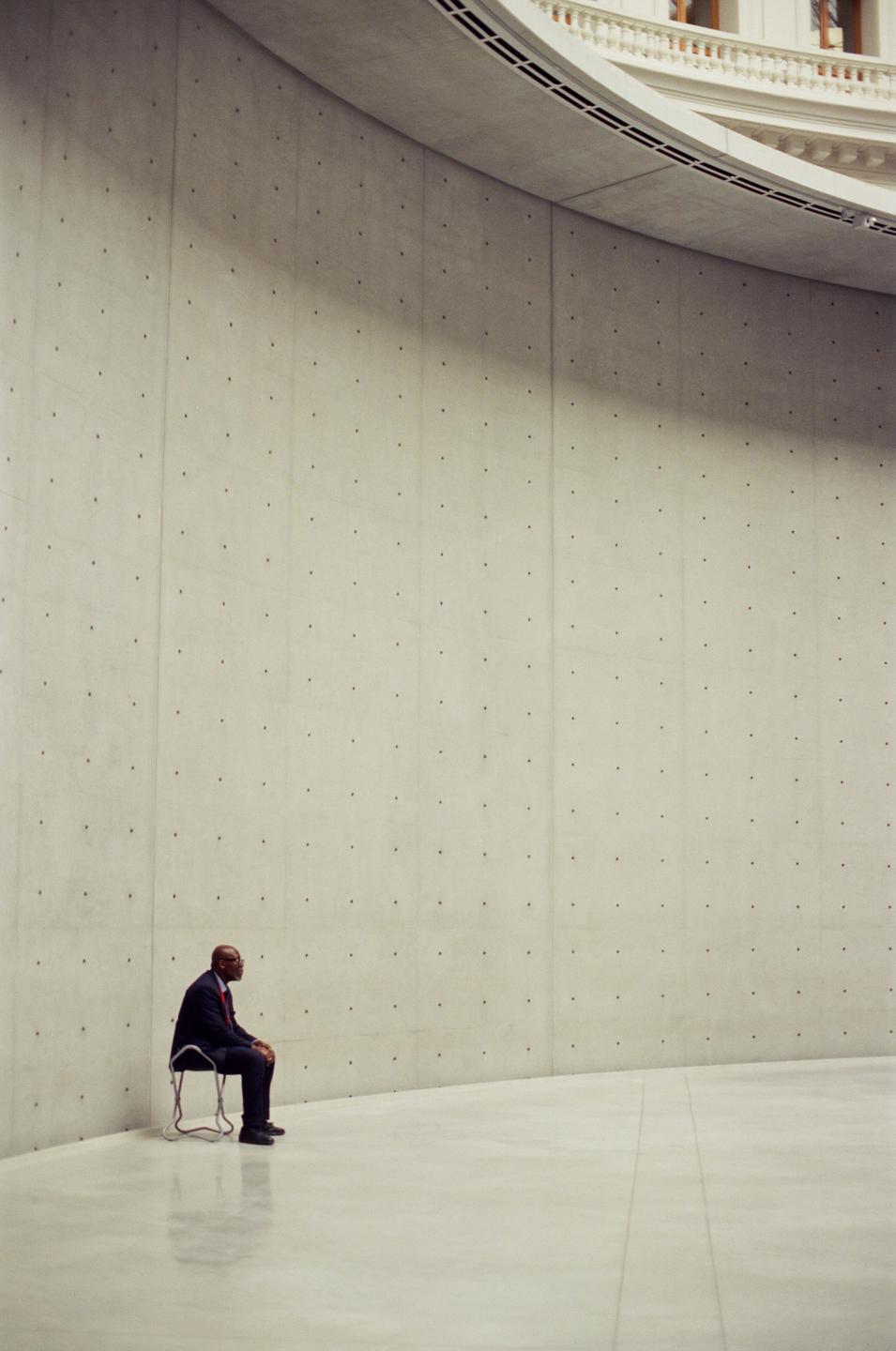 A man in a suit sits on a chair in a large, empty concrete room with a curved wall.