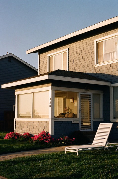 A house with gray shingles and a white lounge chair on the lawn is illuminated by golden hour light.