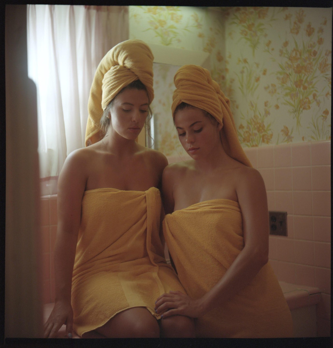 Two women wrapped in yellow towels sit together in a bathroom with floral wallpaper.