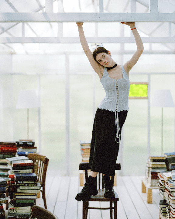 A woman stands on a chair, holding onto a white beam, surrounded by stacks of books in a bright room.