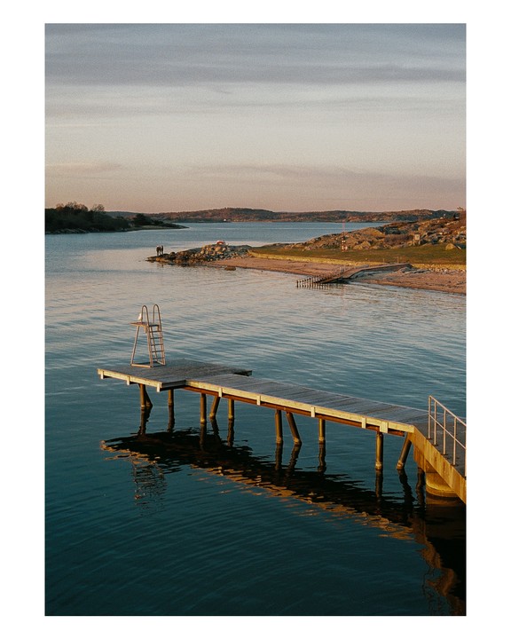 A long wooden pier with a diving board extends into a calm body of water, with a distant shoreline and two people visible.