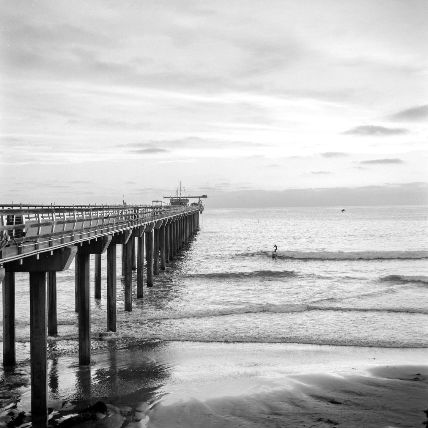 A surfer rides a wave near a long pier extending into the ocean under a cloudy sky.