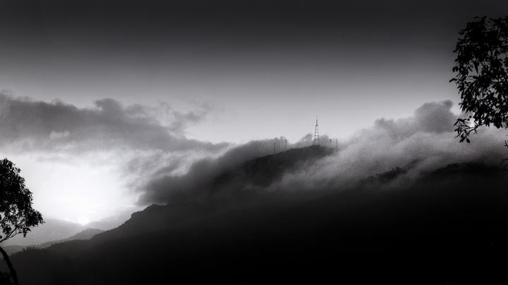 A mountain range is covered in fog with communication towers visible on the peaks and a tree branch in the foreground.