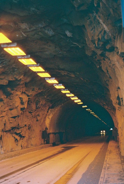 A long, dark tunnel is illuminated by a line of bright yellow lights on the ceiling and two distant headlights.
