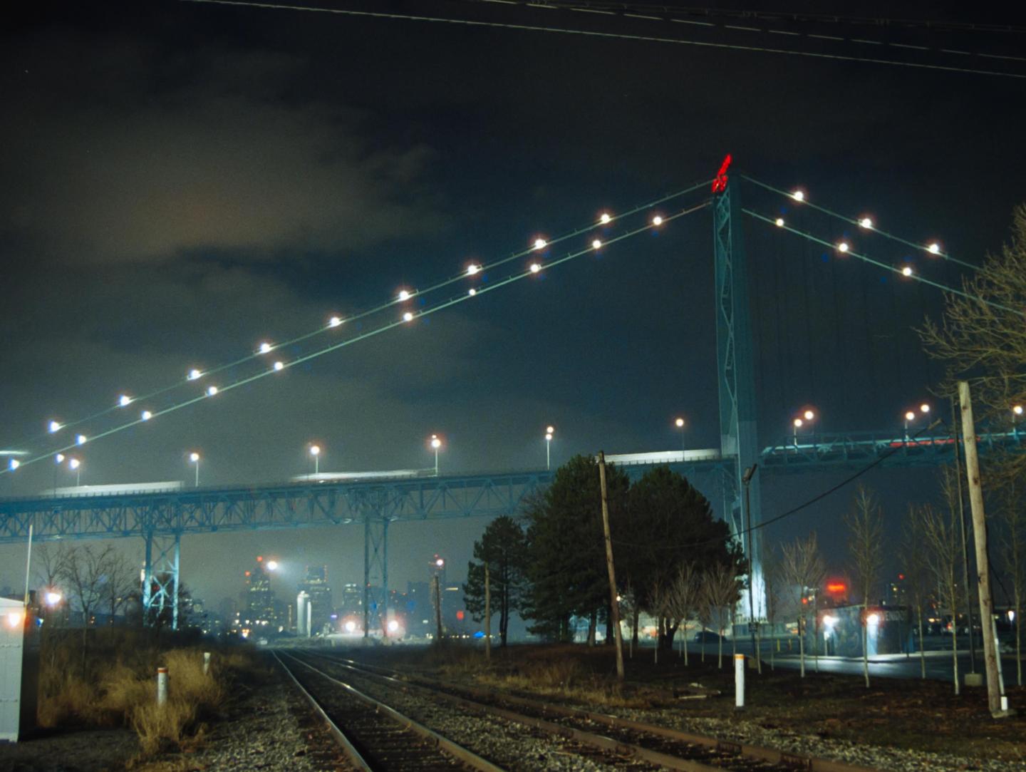 A suspension bridge with lights is visible over railroad tracks and trees at night.