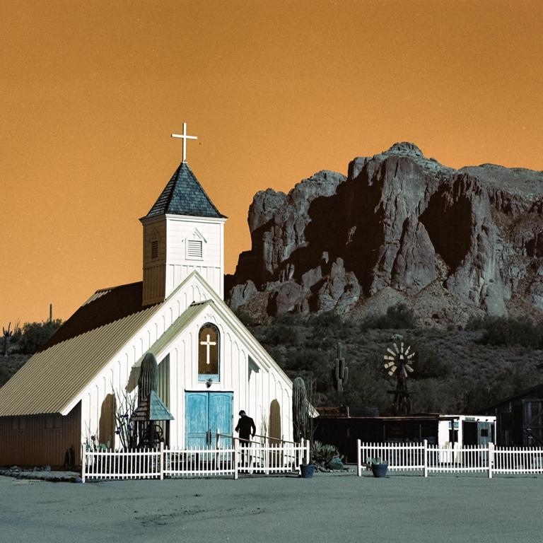 A white church with a steeple and blue doors stands in a desert landscape with mountains in the background.