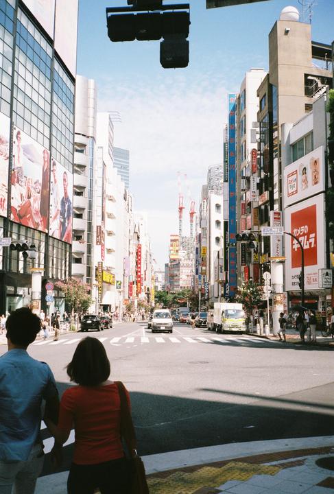 A couple stands at a crosswalk looking down a busy street lined with tall buildings and advertisements.