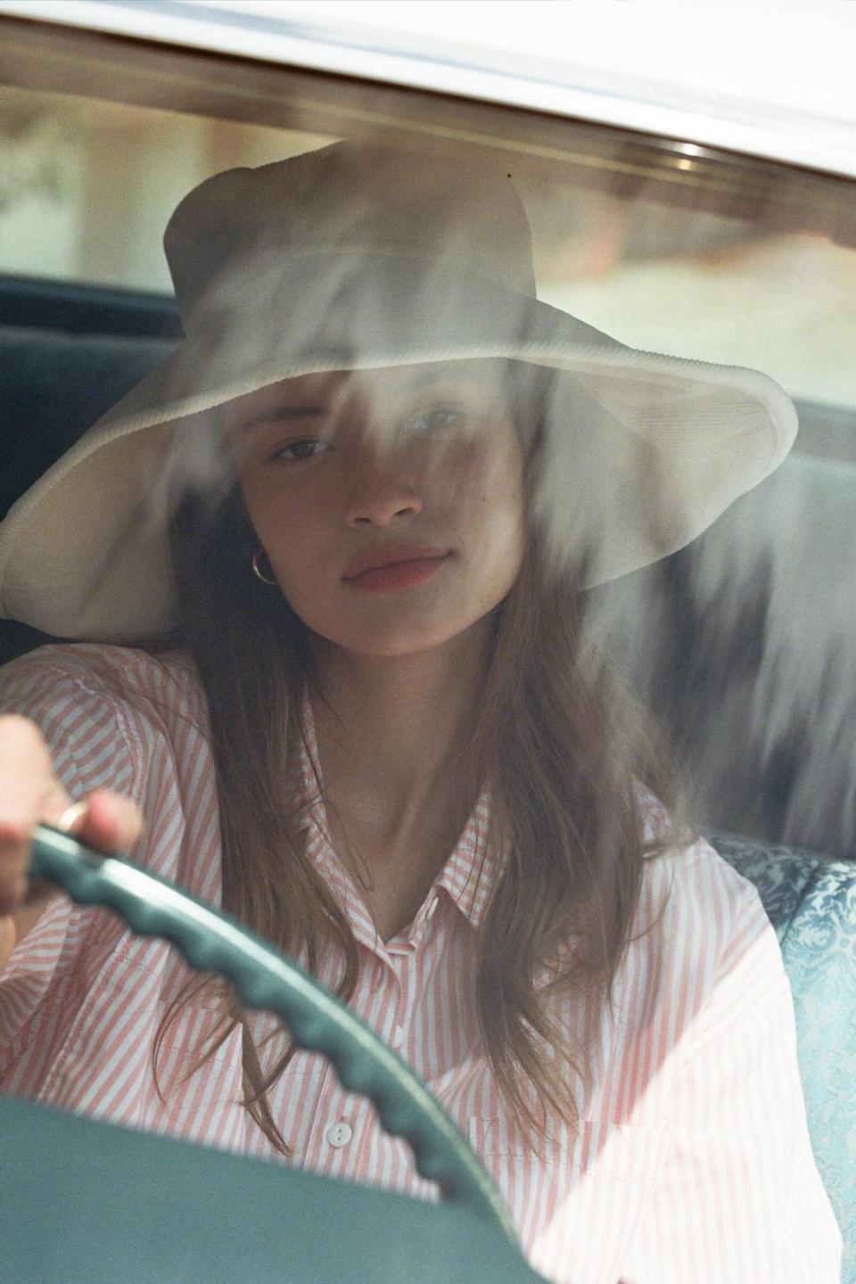 A woman wearing a wide-brimmed hat and a striped shirt is seen through a car windshield, with her hand on the steering wheel.